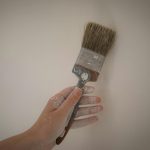 A hand holds a paintbrush with white paint against a plain wall, showcasing DIY home improvement.